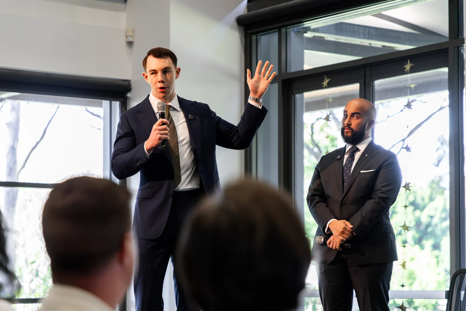 A speaker holding a microphone raises one hand beside another speaker.
